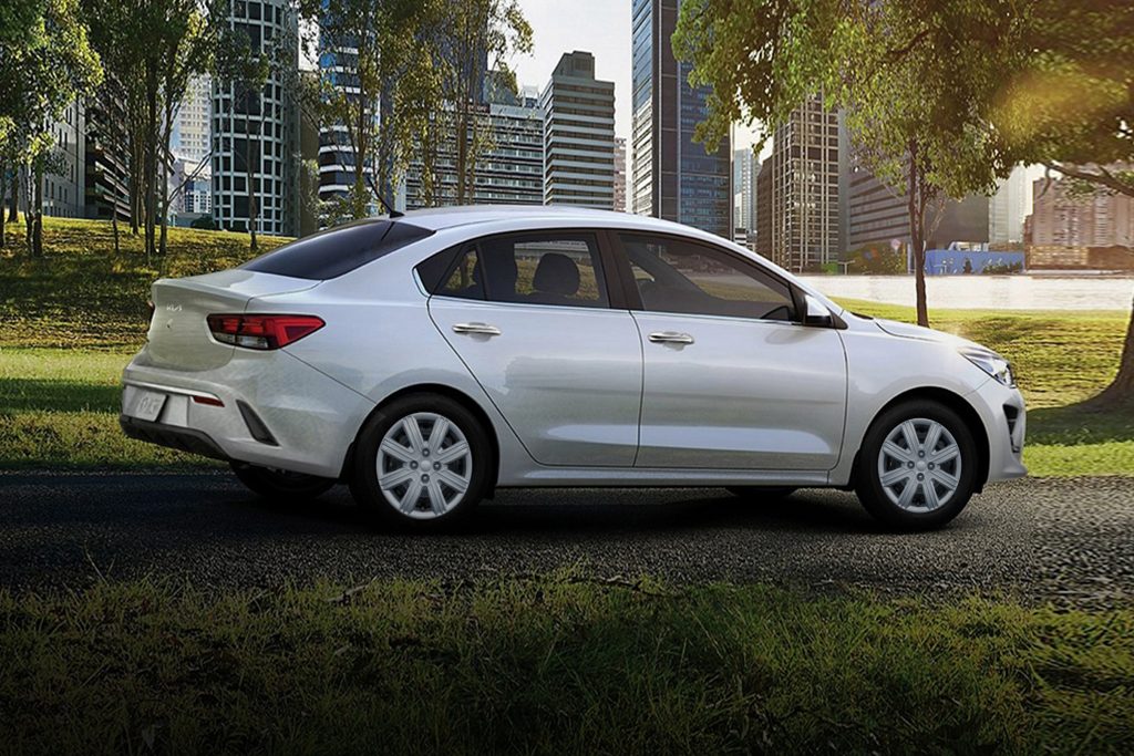 Profile view of a white 2022 Kia Rio parked in a park with buildings in the background. | Kia dealer in Bentonville, AR.