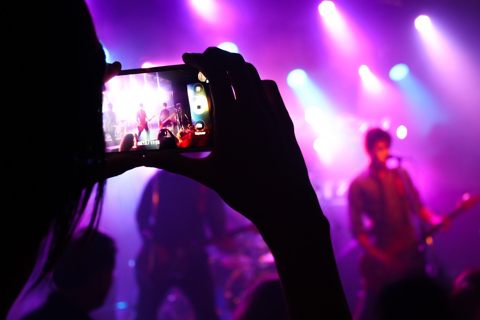A girl holding up a camera recording a band on stage performing. | Theaters and live performances around Bentonville, AR.