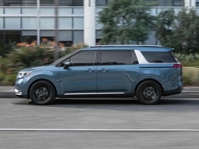 Profile view of a blue 2023 Kia Carnival being driven on the road with a building in the background. | Kia dealer in Bentonville, AR.